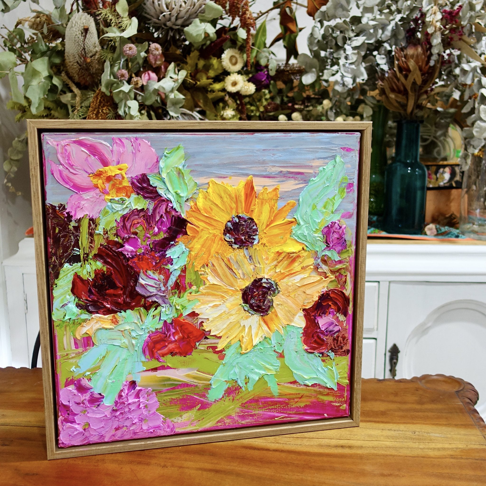  Sunshine is an original painting of yellow, hot pink and red flowers with pale green/blue leaves, acrylic on canvas, 43cm x 43cm. Shown in situ on a timber table in front of vases of flowers.