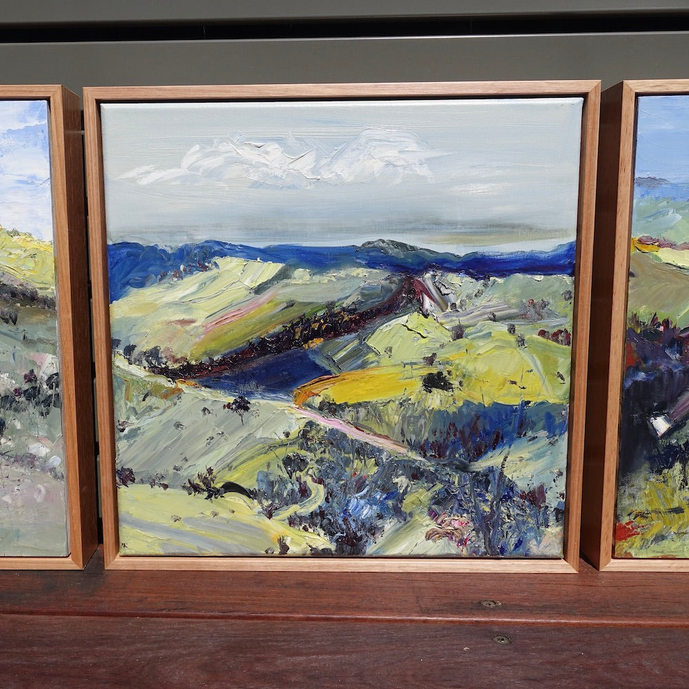 Abstract oil painting of a country landscape with steep hills and lots of colour, yellow, pink, green and blue against a pale blue sky with white clouds. Next to matching paintings.