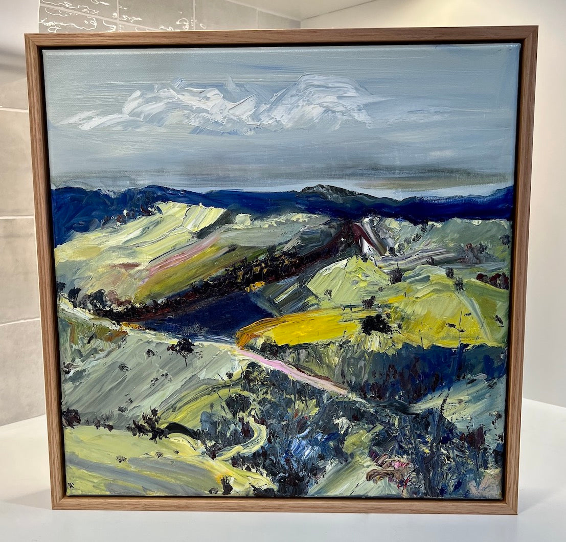 Abstract oil painting of a country landscape with steep hills and lots of colour, yellow, pink, green and blue against a pale blue sky with white clouds. Framed view.