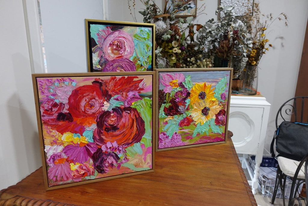 Sunshine is an original painting of yellow, hot pink and red flowers with pale green/blue leaves, acrylic on canvas, 43cm x 43cm. Shown in situ on a wooden table with two other paintings.