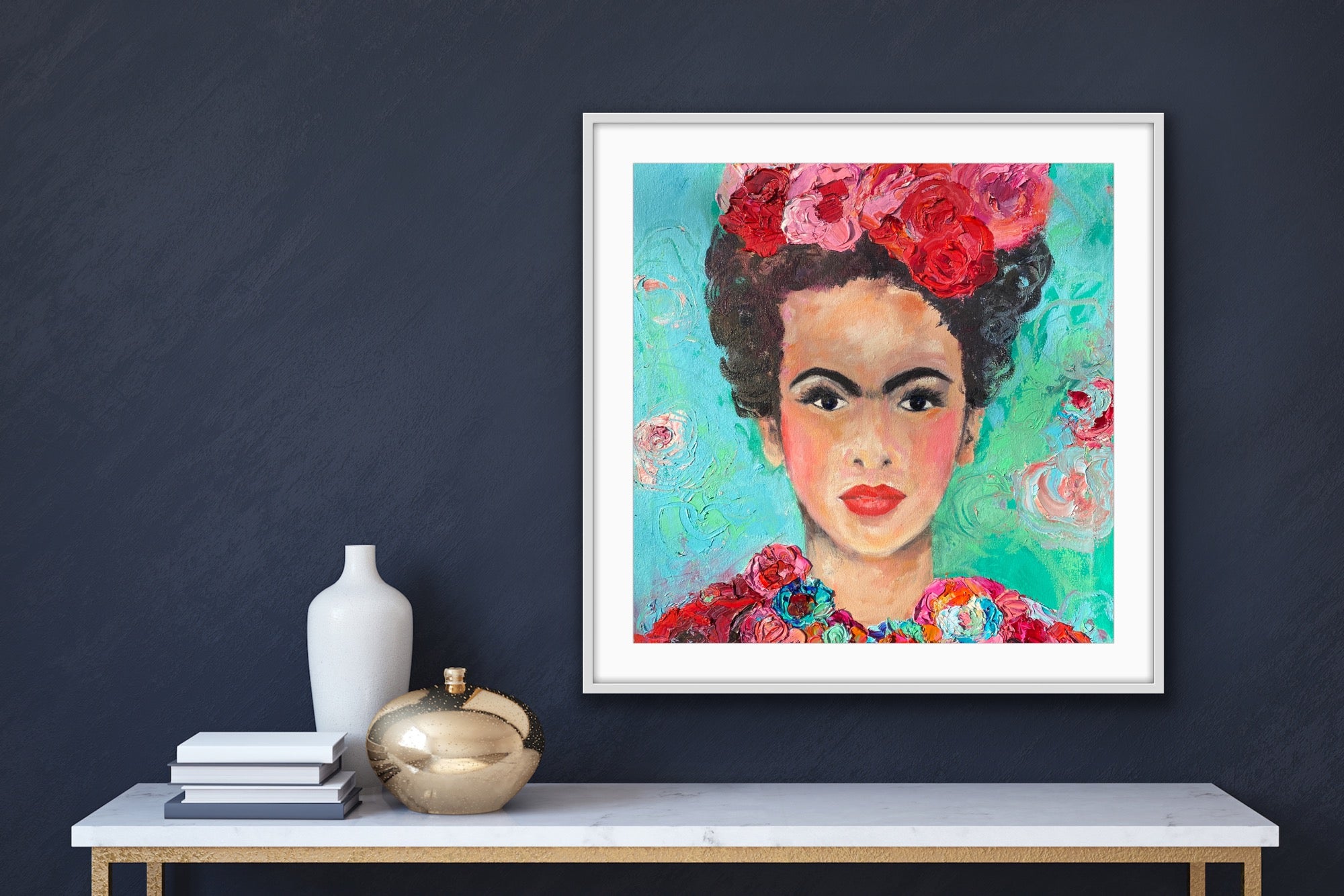 A beautiful and colourful print of Frida Kahlo with red and pink flowers in Frida's hair against a turquoise and aqua background. Framed in white against a dark grey wall.