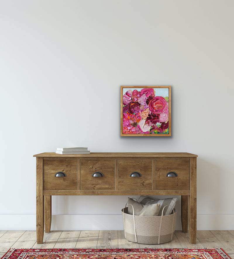 An original painting of a gorgeous bunch of florals in all shades of pink, entwined with gold thread on a pale blue background, shown on a white wall.