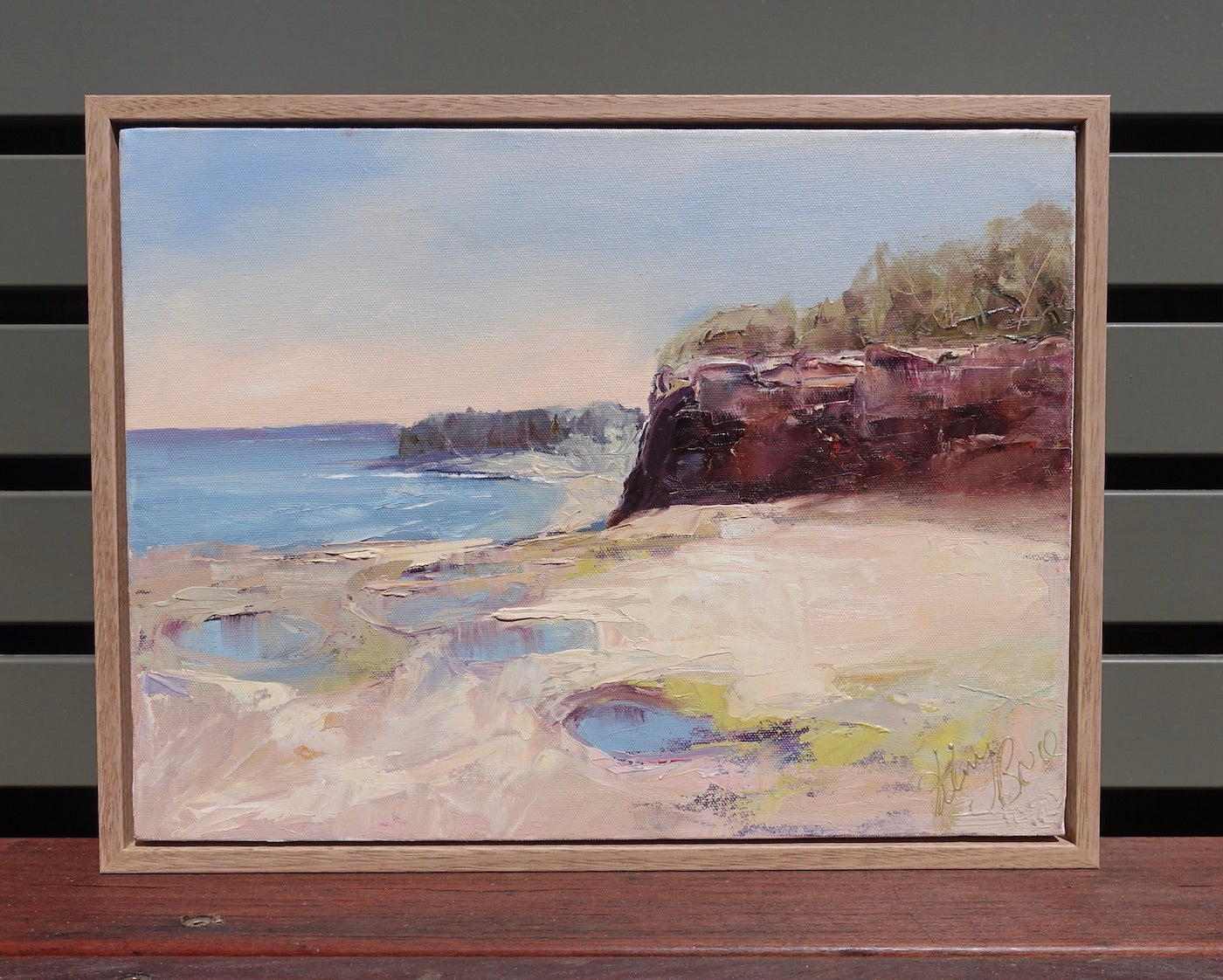 Ocean and rock pools in soft pastel colours with reddish brown cliffs. Framed view.